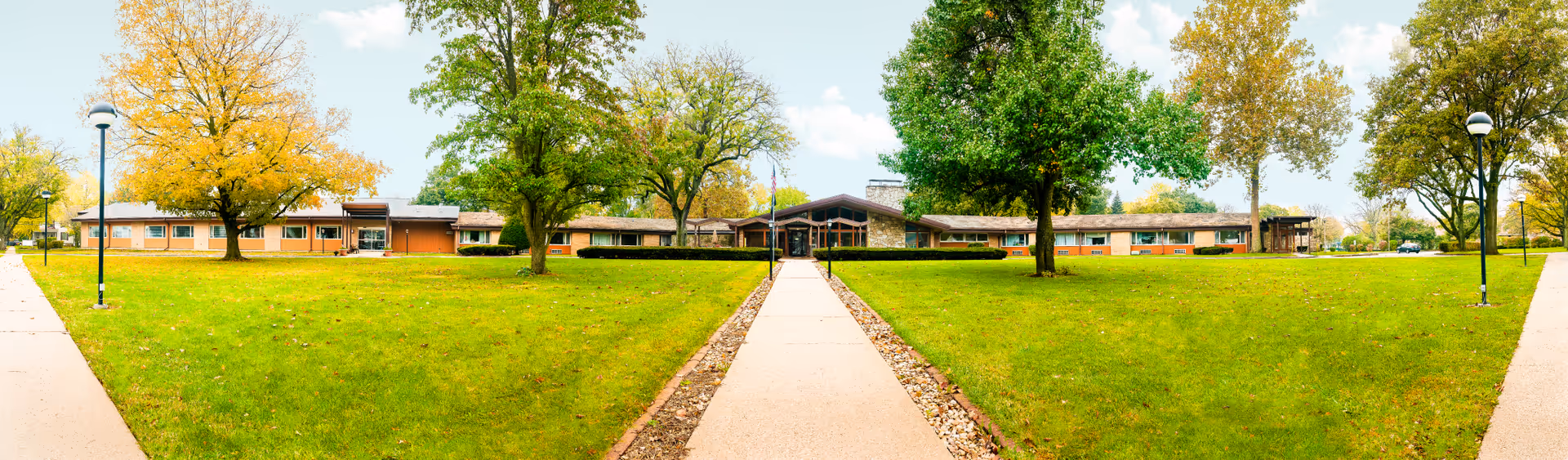 Wide panoramic view of a single-story healthcare facility building with a stone and wood exterior, surrounded by green lawns and trees with autumn foliage. A concrete walkway leads straight to the main entrance, flanked by lamp posts on either side. The sky is partly cloudy.