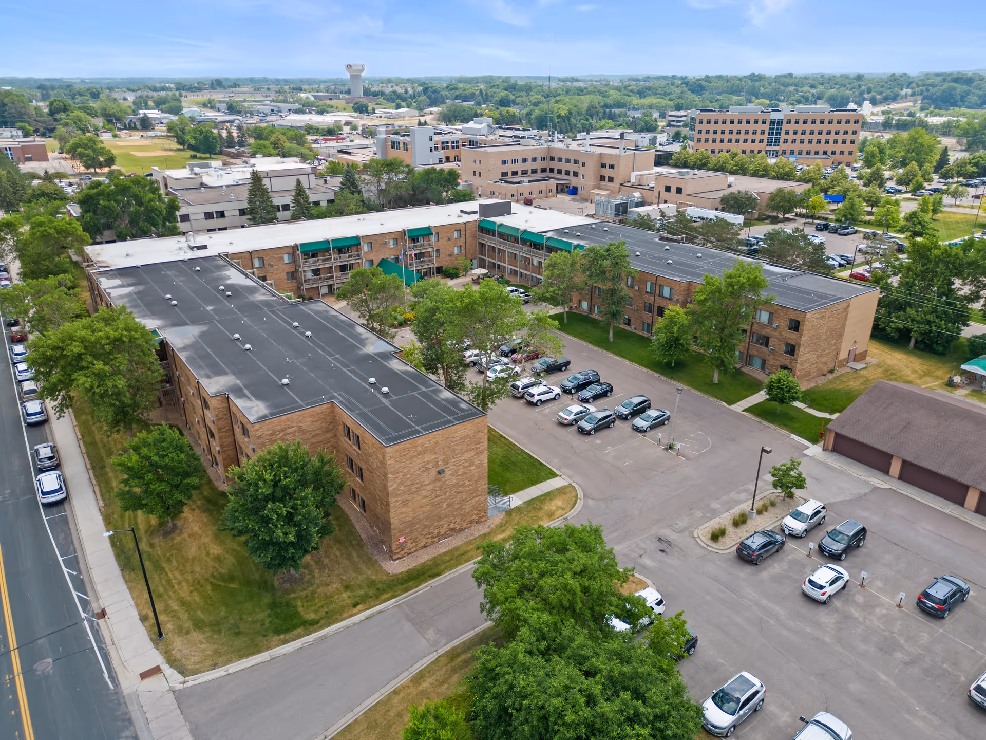 Aerial view of Good Samaritan Society - Waconia facility showing a large, multi-story brick building with a flat roof surrounded by trees and parking lots with several parked cars. The surrounding area includes other buildings, roads, and greenery under a clear sky.