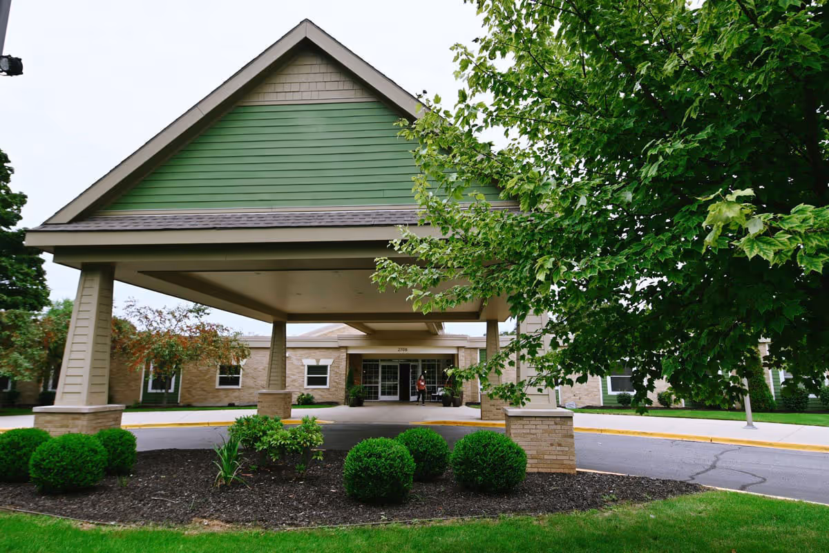 Covered entrance canopy and porte-cochère at the front of a low-rise senior living building with shrubs and a large tree.