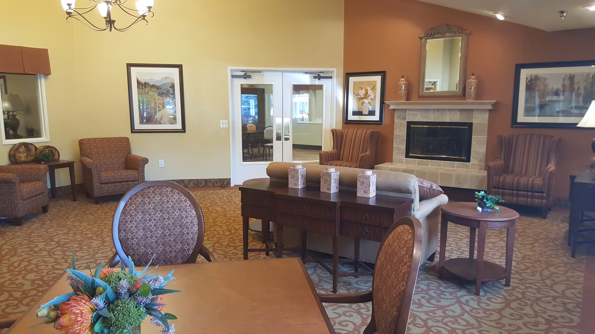 A cozy living room area in a senior living facility with patterned carpet, several upholstered armchairs, a sofa, a wooden table with decorative items, a fireplace with a mirror and vases above it, framed artwork on the walls, and a chandelier hanging from the ceiling.