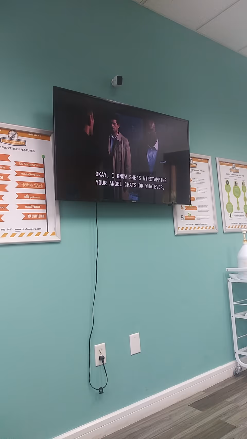 Wall-mounted TV shows subtitles; teal walls, posters, and power cord visible.