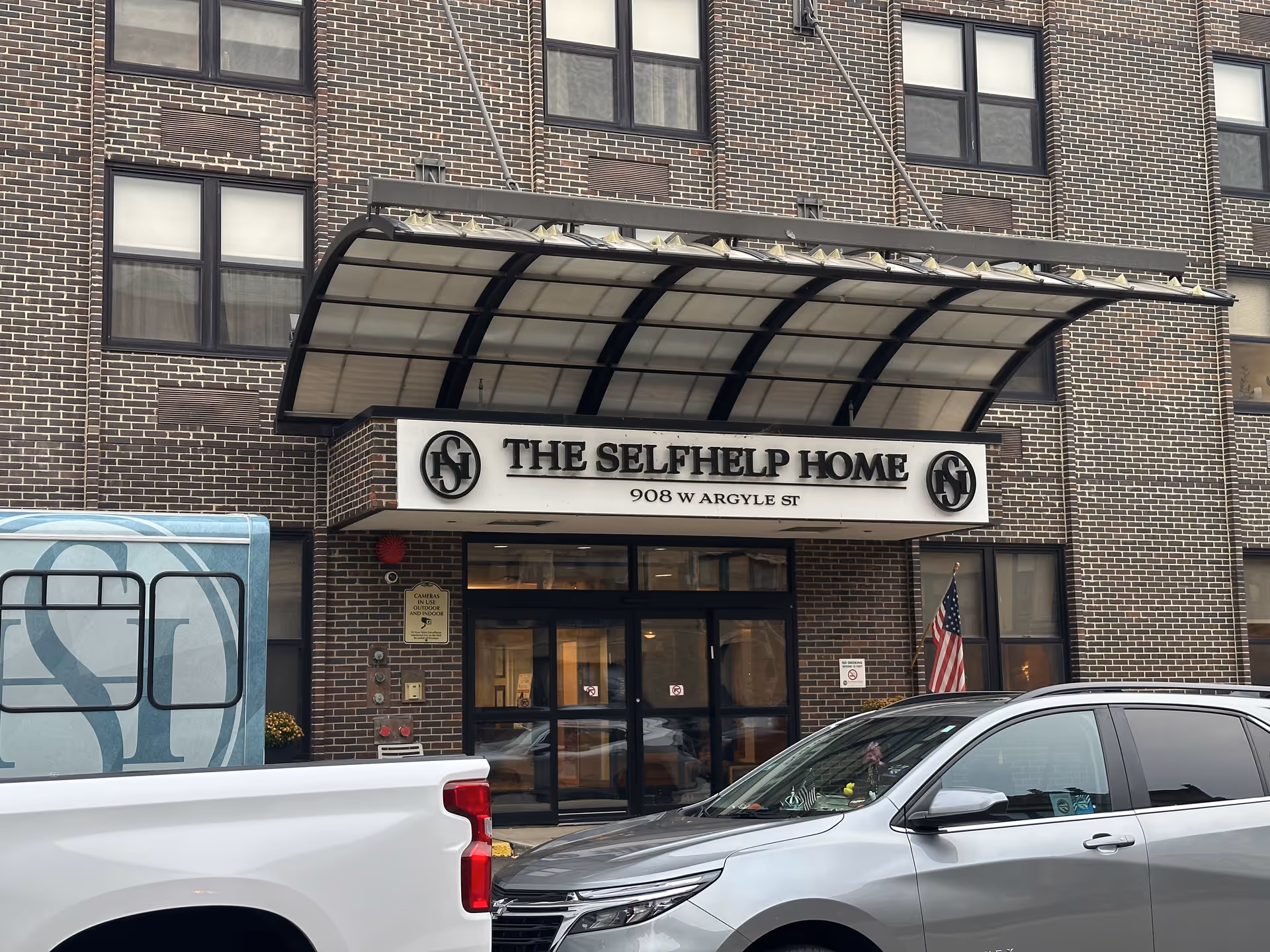 Exterior front entrance of The Selfhelp Home Senior Living Community building with a large awning above the doorway, brick facade, and two vehicles parked in front.