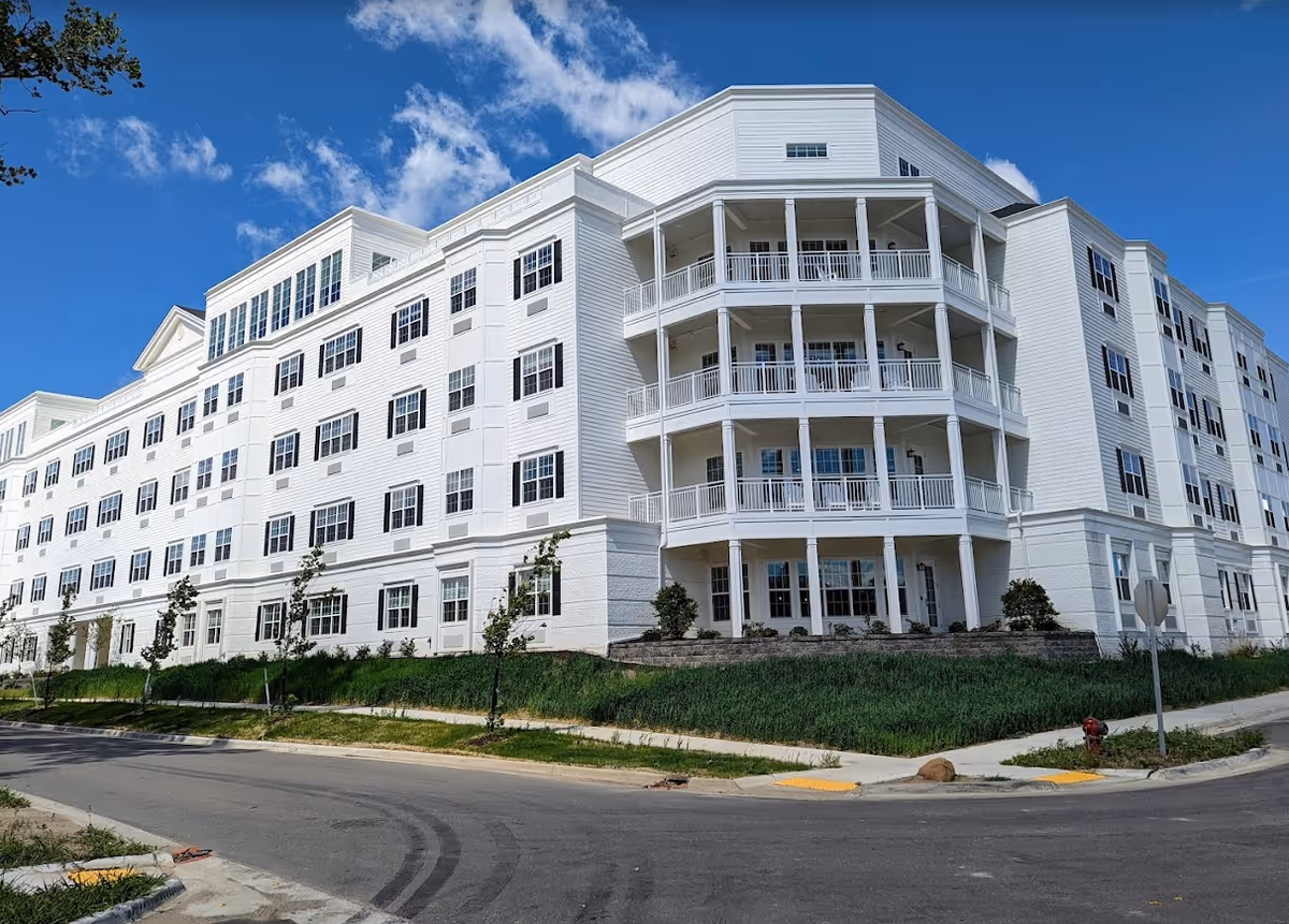 Large white multi-story residential building with stacked wraparound balconies, many windows, and landscaped grounds under a blue sky.