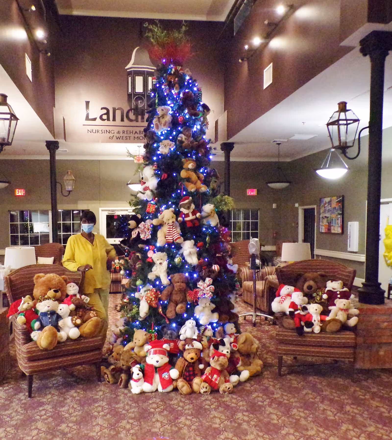 A decorated Christmas tree adorned with numerous teddy bears and blue lights stands in the center of a cozy room. Two armchairs on either side of the tree are also filled with teddy bears. A person wearing a yellow outfit and a face mask stands to the left of the tree. The room has warm lighting, patterned carpet, and visible signage for Landmark Nursing & Rehabilitation Center of West Monroe, LLC on the wall behind the tree.
