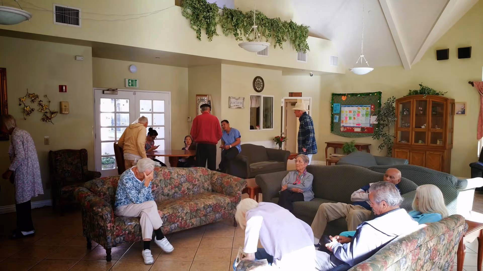A common living area in a senior living facility with several elderly residents sitting on floral and solid-colored couches and chairs. Some people are engaged in conversation while others sit quietly. The room has high ceilings, hanging light fixtures, plants, a bulletin board, and a wooden cabinet. There is an exit door with windows letting in natural light.