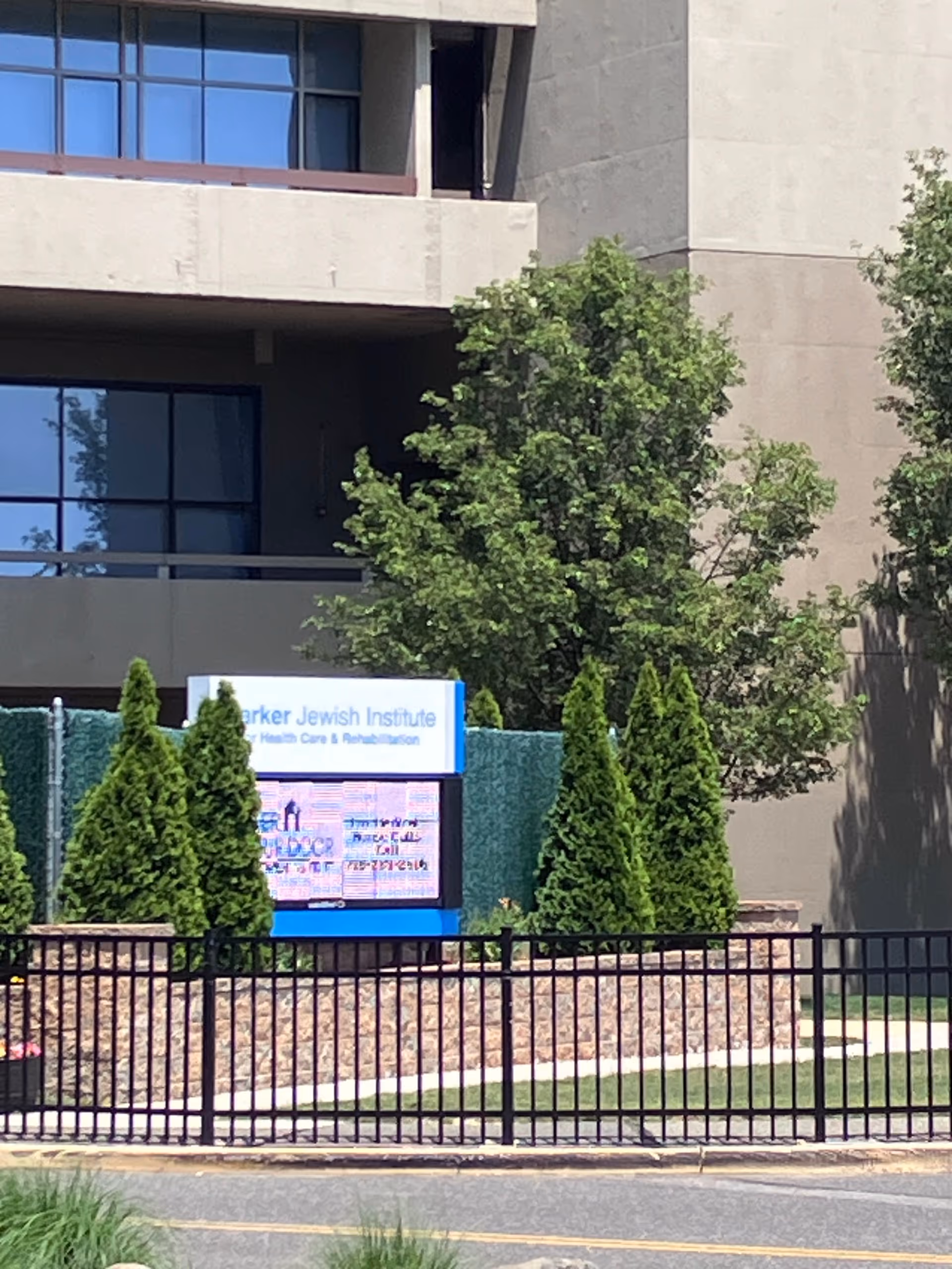 Front exterior of Parker Jewish Institute with sign, evergreen shrubs, and a black fence.