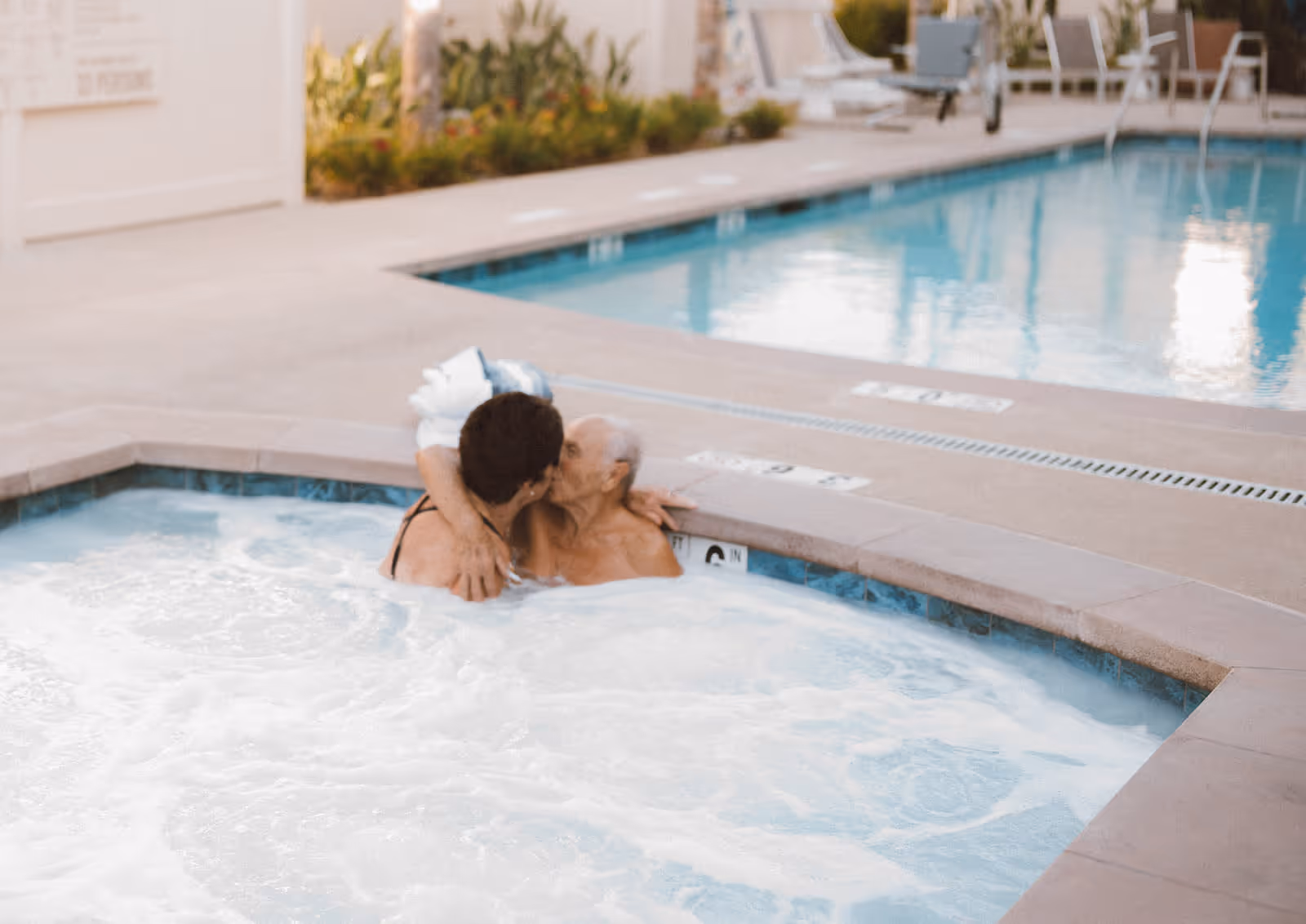 Two elderly people embracing and kissing while sitting in a bubbling hot tub next to a swimming pool at an outdoor facility.