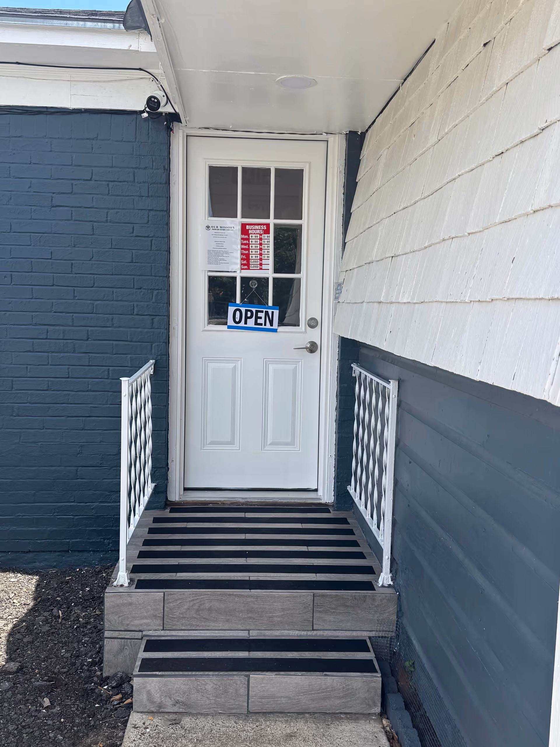 White door with OPEN sign, blue brick exterior, white railings, and wooden steps.
