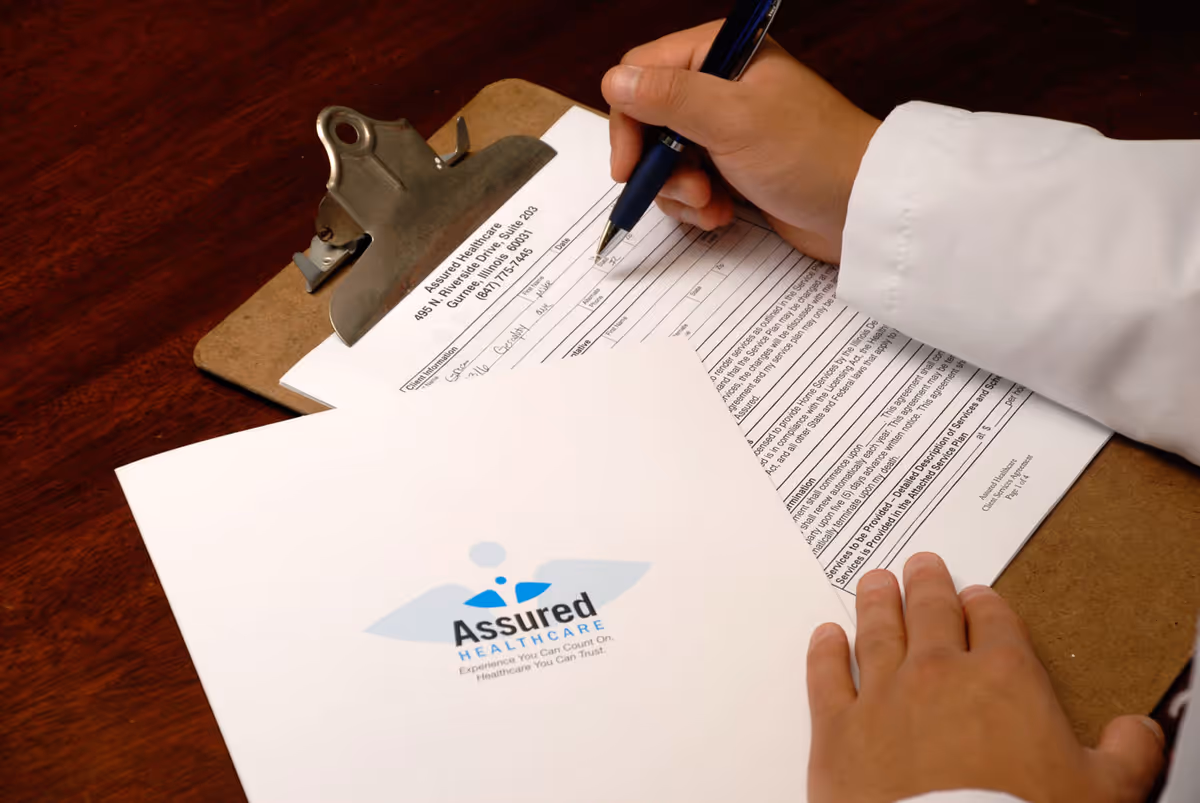 A clinician signs forms at a clipboard with an Assured Healthcare Staffing document.