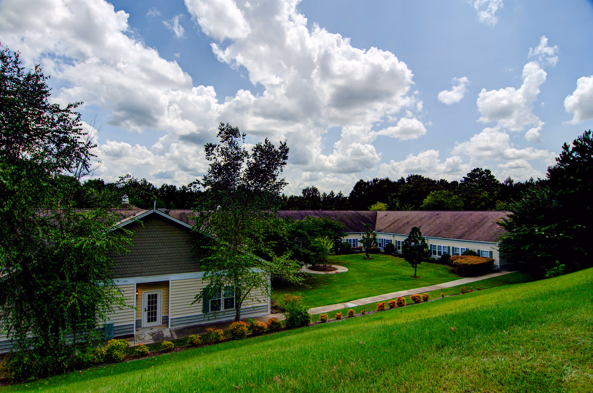 Exterior view of a single-story building with beige siding and a brown roof, surrounded by green grass, trees, and shrubs under a partly cloudy sky.