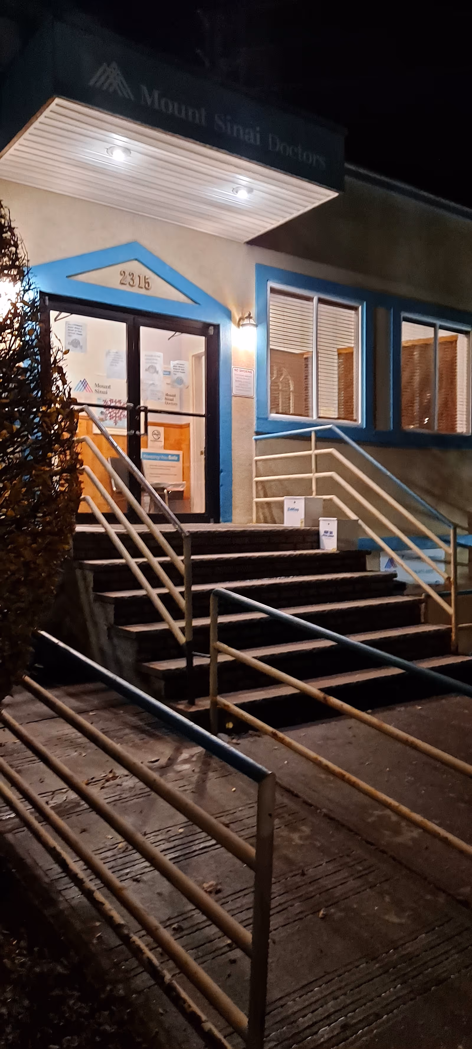 Night exterior view of Mount Sinai Doctors entrance with stairs, rails, and blue-trimmed facade.