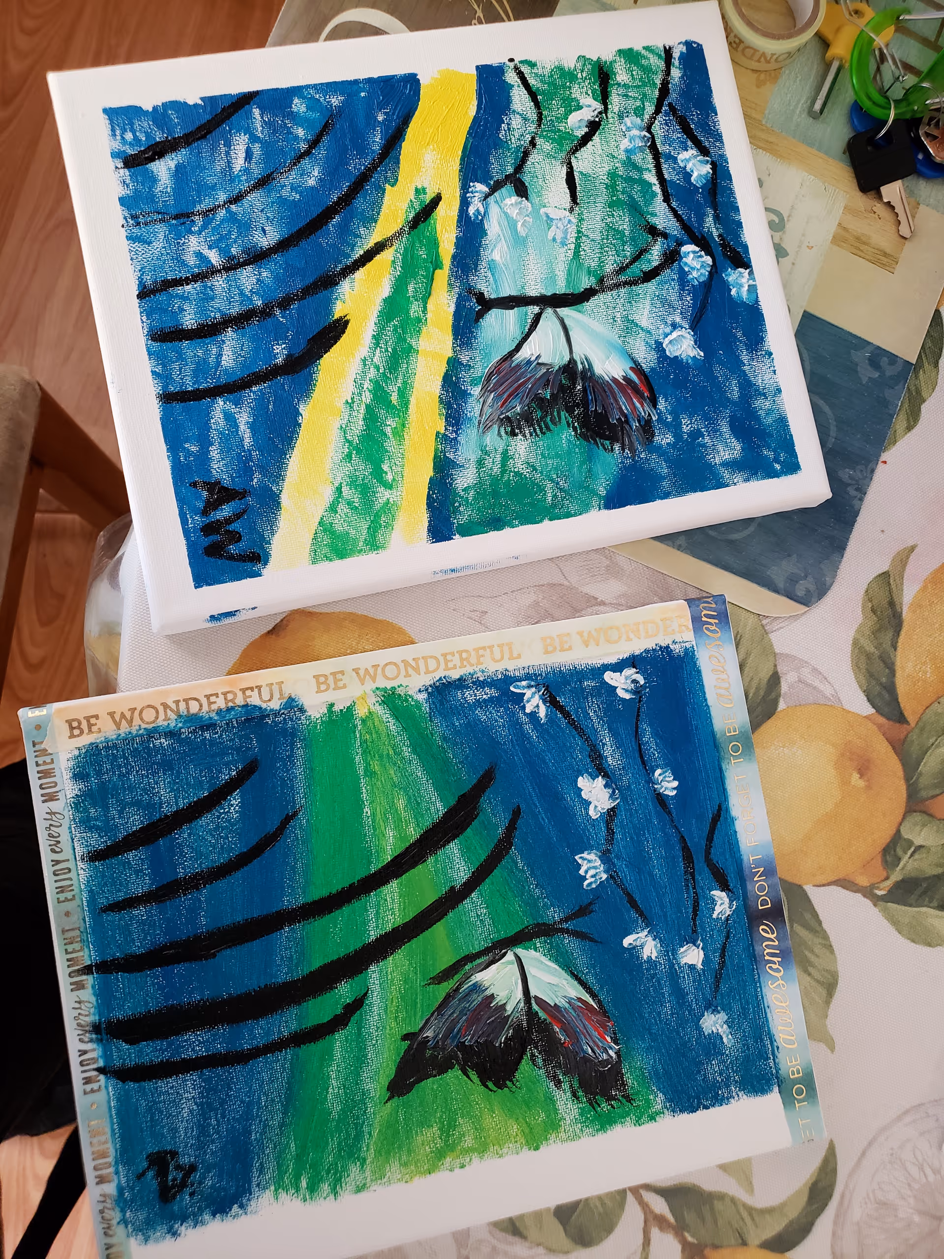 Two colorful abstract paintings on a table with a floral tablecloth inside a room