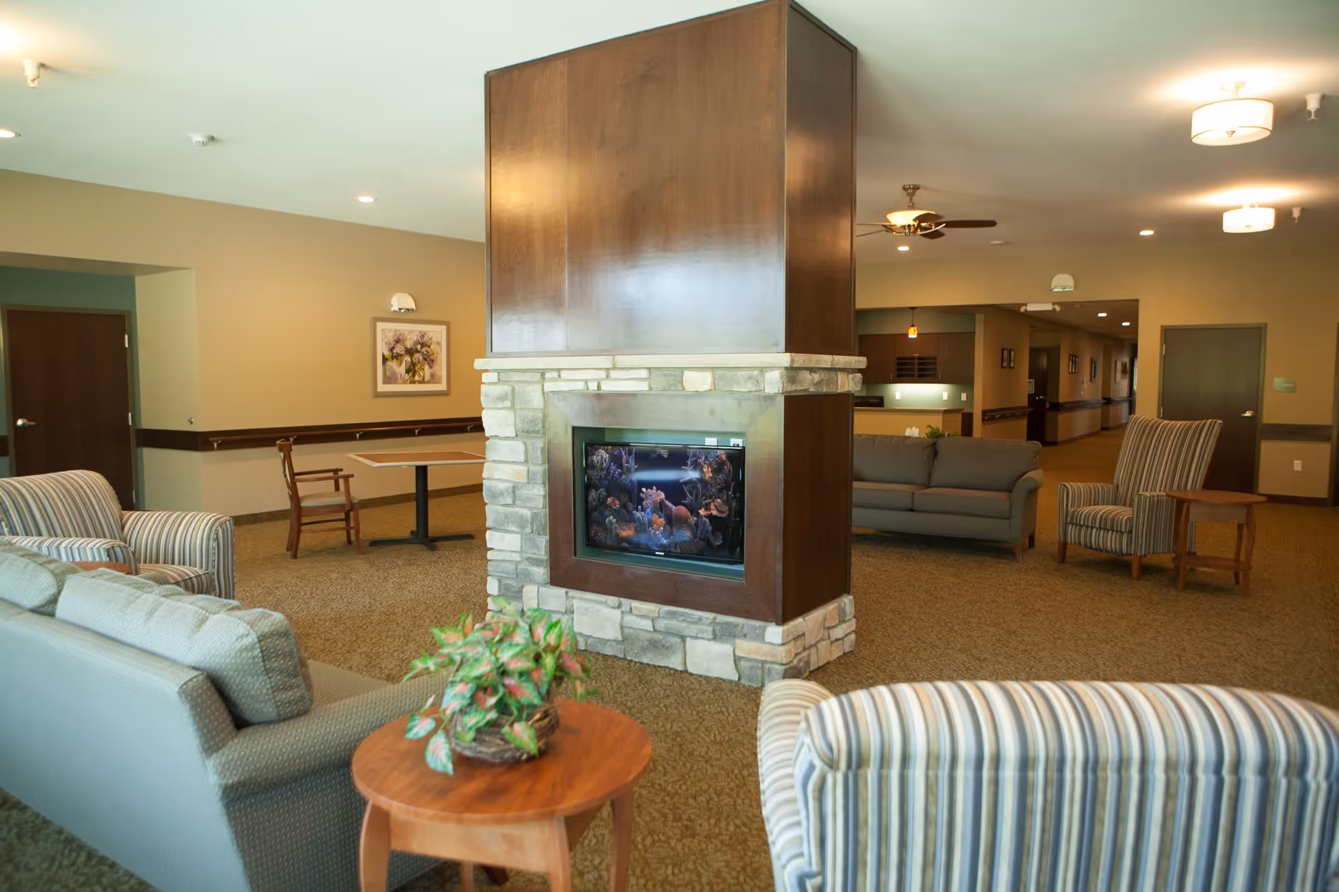 A cozy living room area in a senior living facility featuring a central stone and wood fireplace with an aquarium inside. The room is furnished with striped and solid sofas and armchairs, a small wooden table with a potted plant, and soft lighting from ceiling fixtures and a ceiling fan. The walls are painted in neutral tones with framed artwork and doors leading to other rooms.