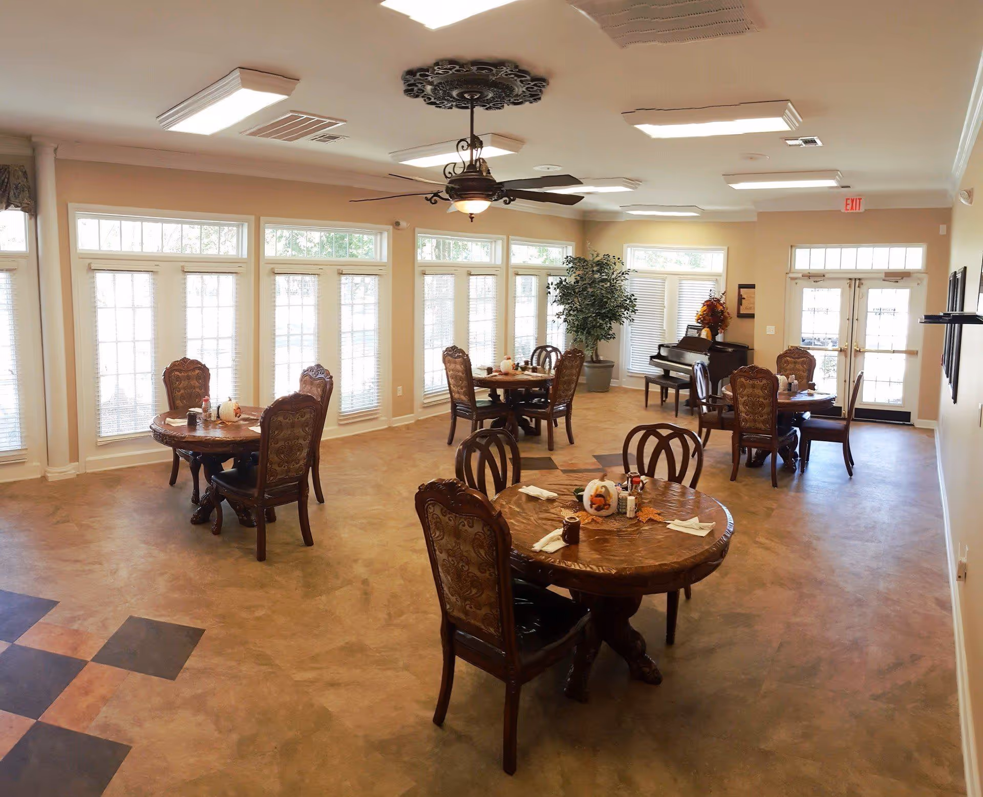 Bright dining/common room with round wooden tables and ornate chairs, large windows, a ceiling fan, and a piano in the background.