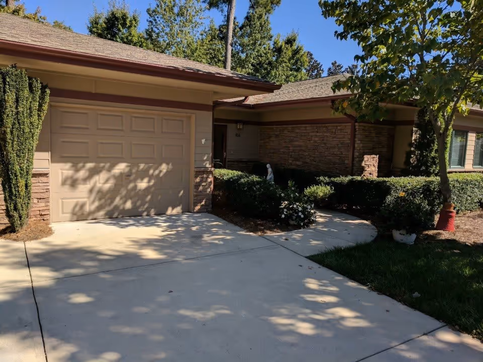 Exterior view of a single-story building with a closed beige garage door, stone and siding walls, surrounded by green shrubs and trees casting shadows on the driveway.
