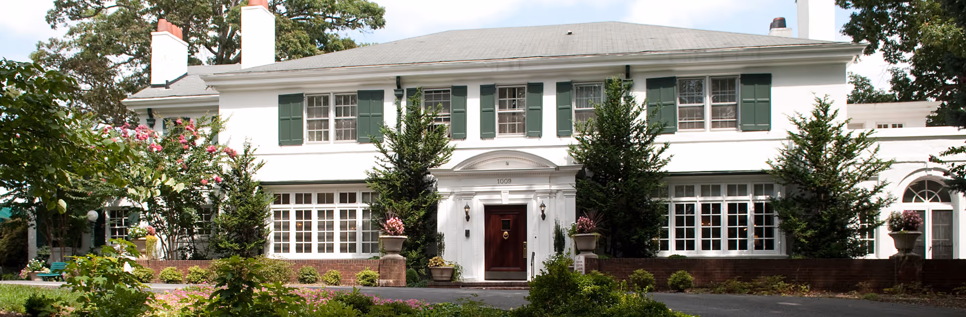 Front exterior of a two-story white colonial-style building with green shutters, a central doorway, and landscaped grounds.
