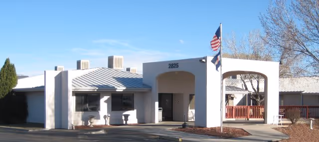White, low-rise senior care facility with archway entrance, flagpole, and blue sky.