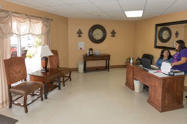 Reception area of Alta Vista Rehabilitation & Healthcare with two women at a wooden desk, a large mirror on the wall, a clock, a small table with a coffee maker, and two upholstered chairs near a window with curtains.