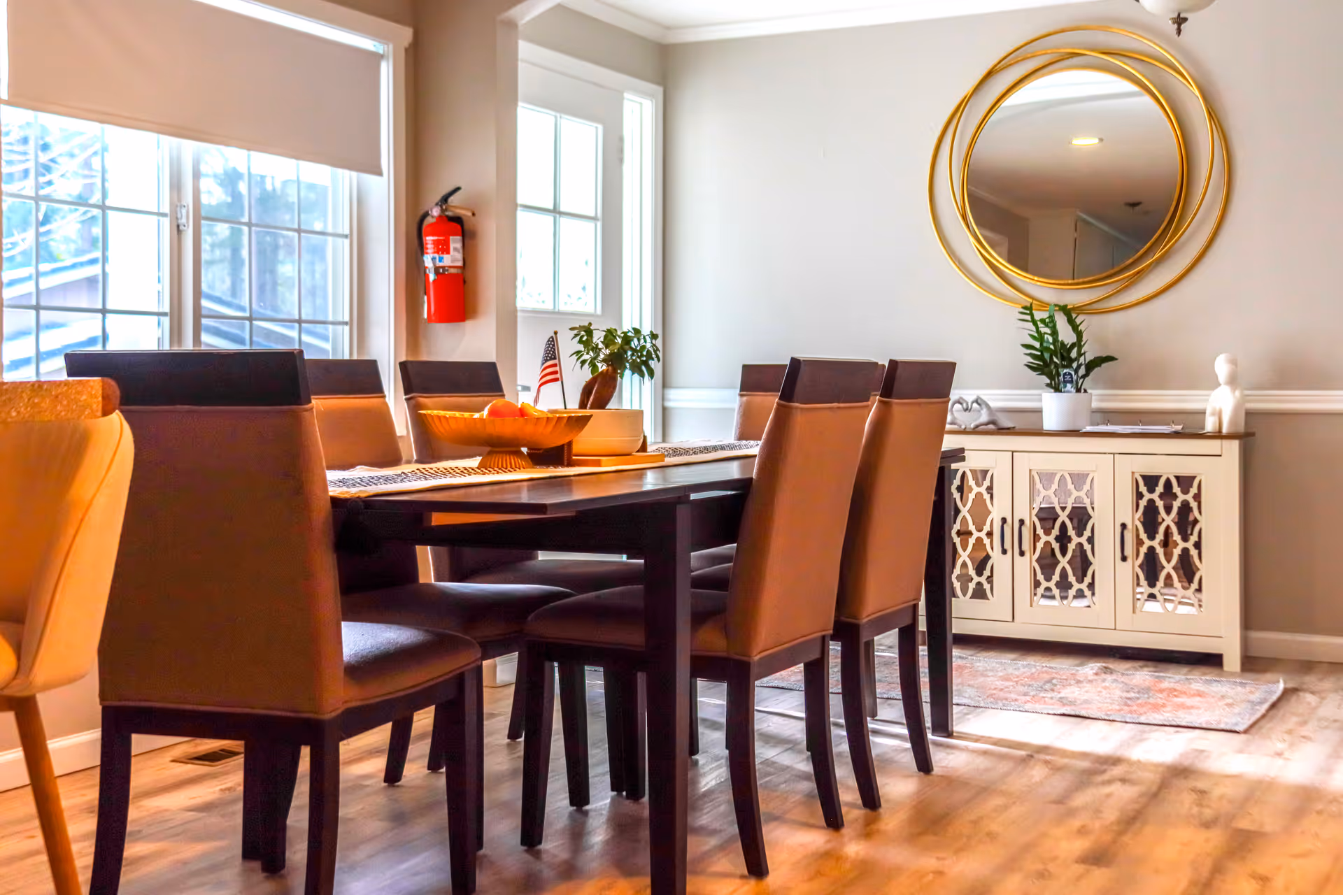 Bright dining room with a dark table, tan chairs, sideboard, and round gold mirror.