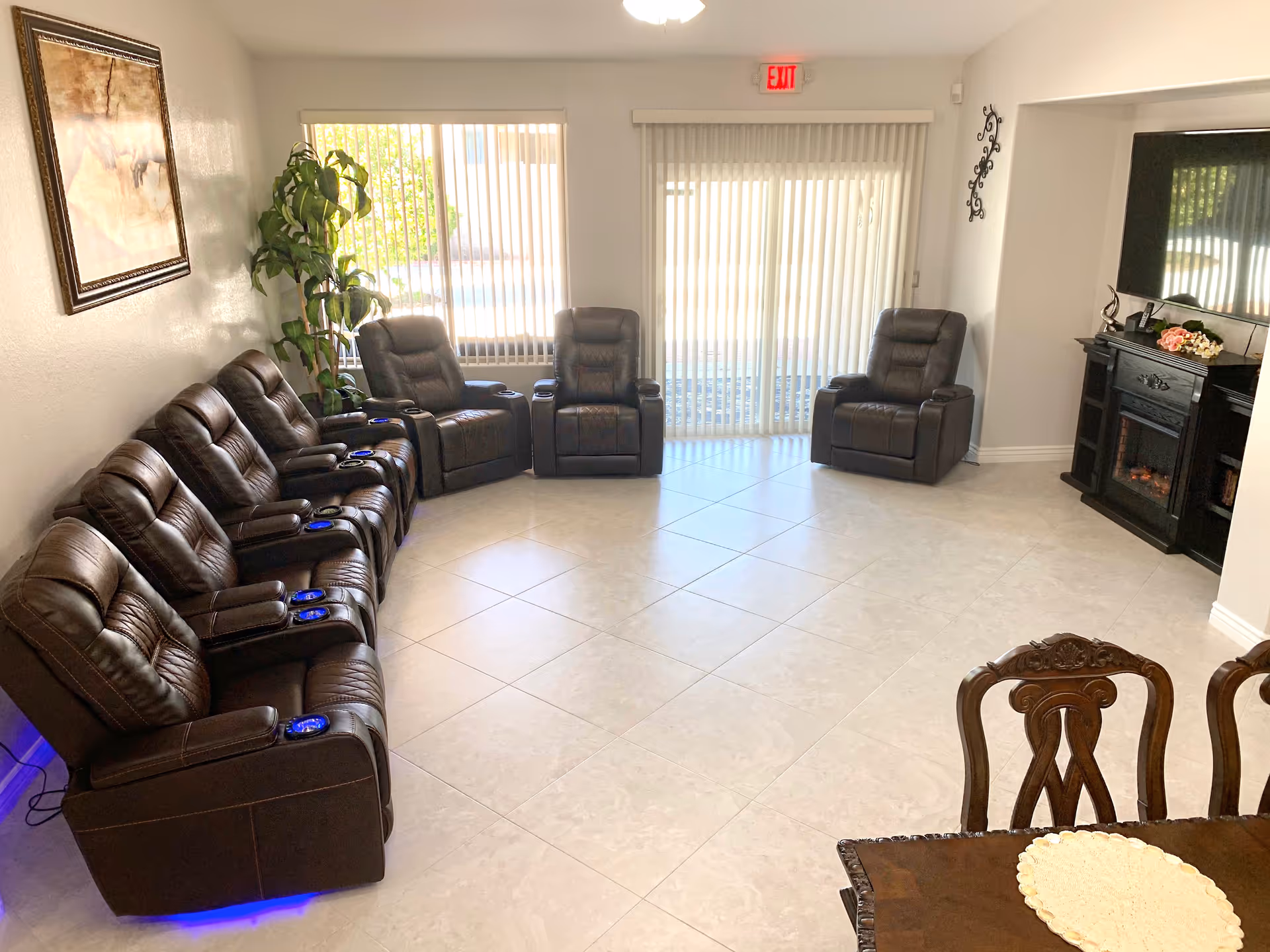 A bright living room with a row of six brown leather recliners along the left wall and three more recliners arranged near large windows with vertical blinds. There is a large flat-screen TV mounted above a black electric fireplace on the right wall. A wooden dining table with ornate chairs is partially visible in the foreground. A potted plant and framed artwork decorate the left wall.