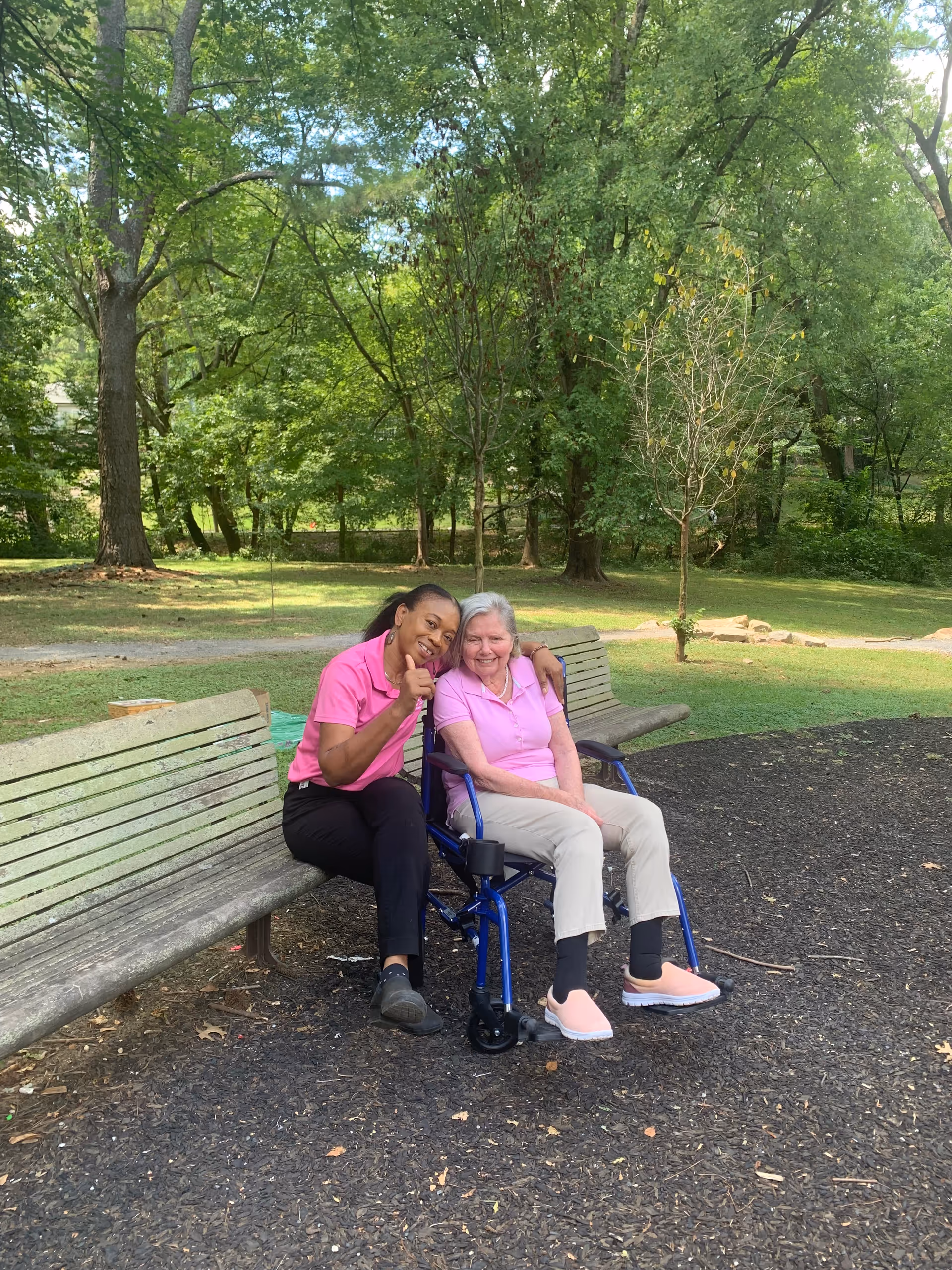 Two people sit on a park bench outdoors; one in a wheelchair beside a caregiver.