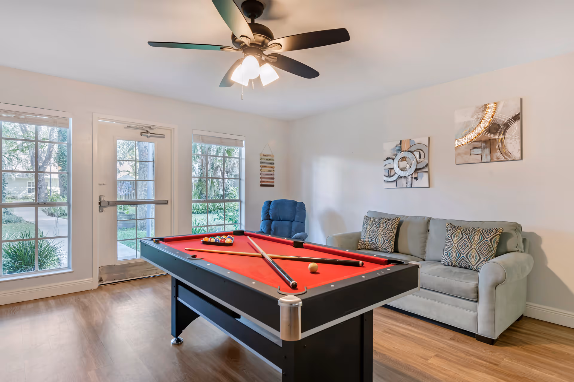 Pool table with red felt in a bright living room with sofa and ceiling fan.