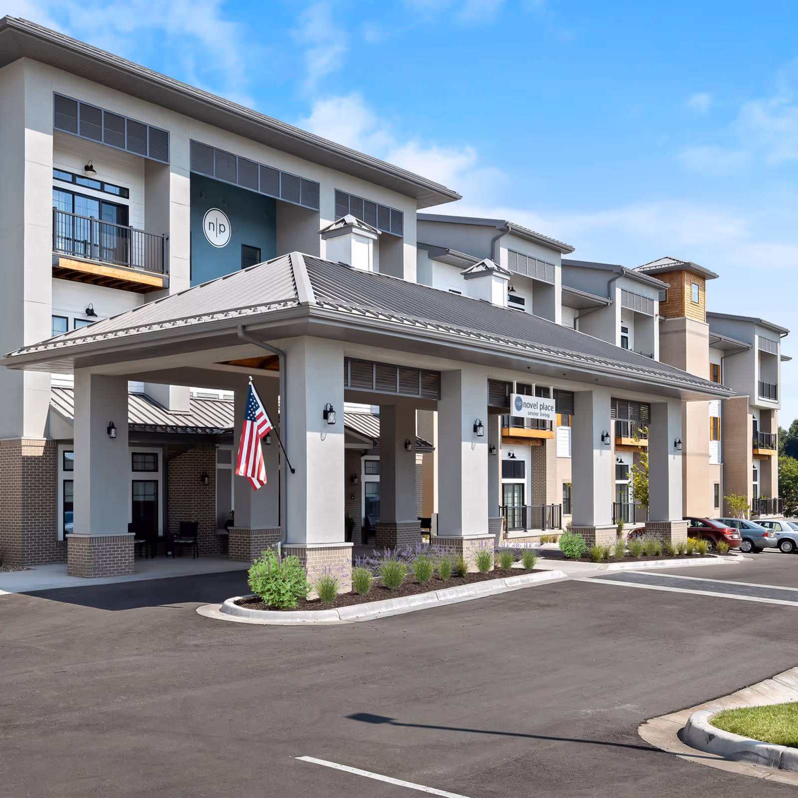Front exterior of Novel Place Senior Living - Blue Springs showing a modern multi-story building with a covered porte-cochere, American flag, landscaping, and parked cars.