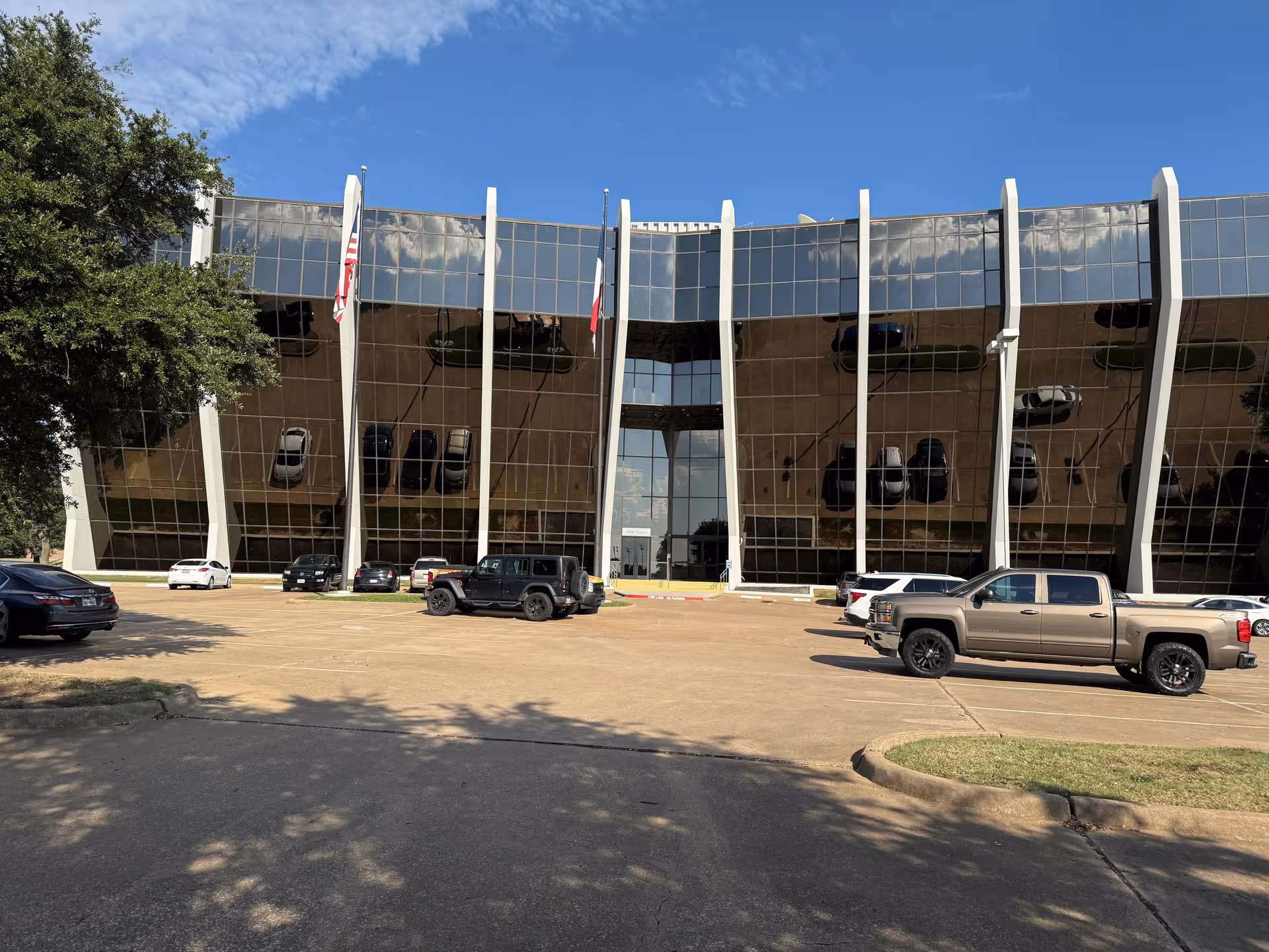 Modern glass-front building with tall vertical columns, flags, and parked vehicles in front.