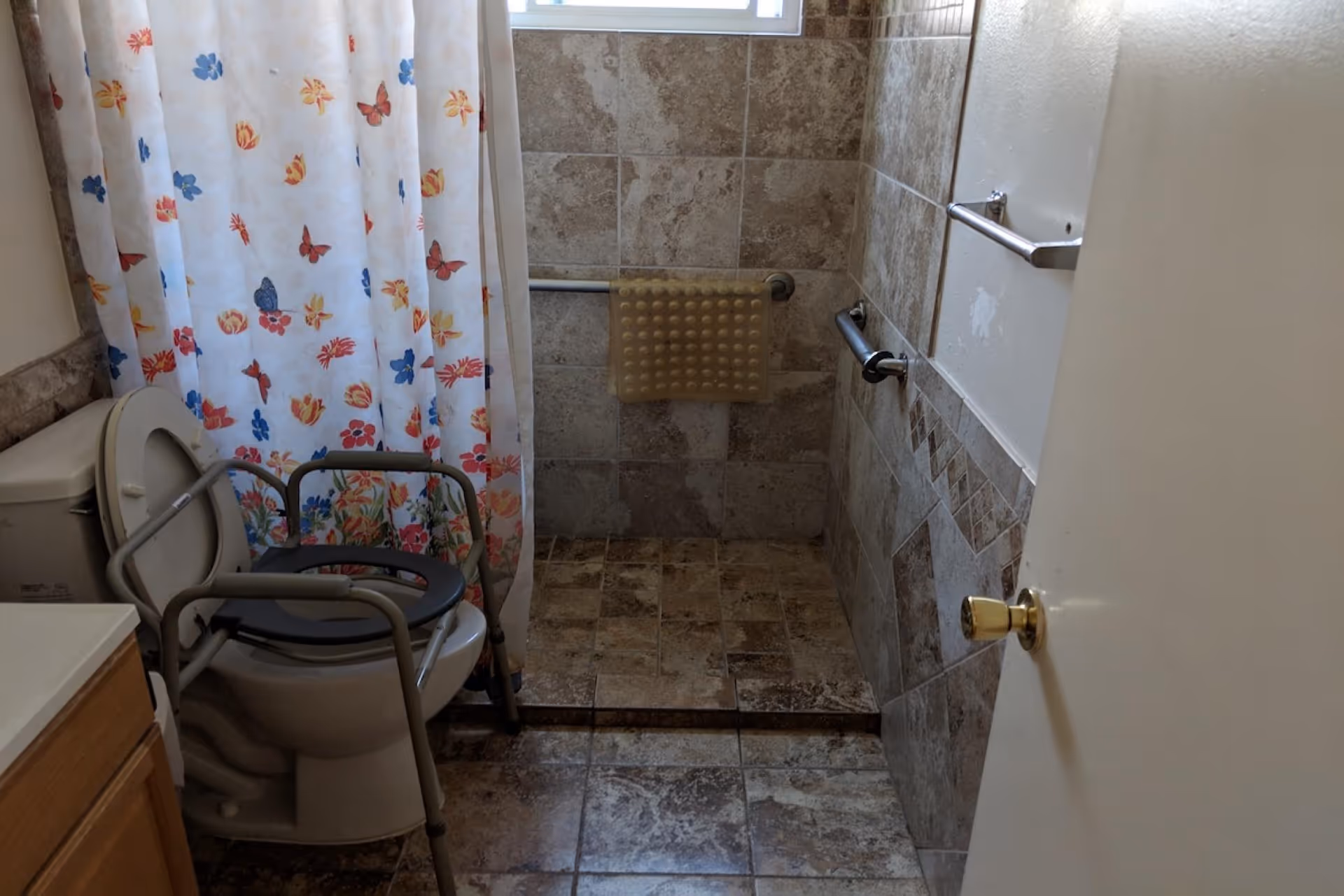 A bathroom with a tiled floor and walls, featuring a toilet with a raised seat and safety frame, a shower area with a floral and butterfly patterned shower curtain, grab bars on the walls, and a textured bath mat hanging on the wall.