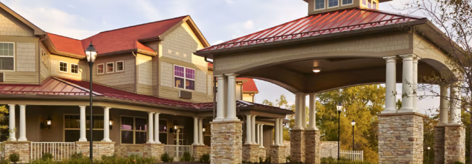 Exterior view of a senior living facility building with a covered entrance supported by stone and white columns, red metal roofs, beige siding, and multiple windows. There are lamp posts and some greenery around the building.