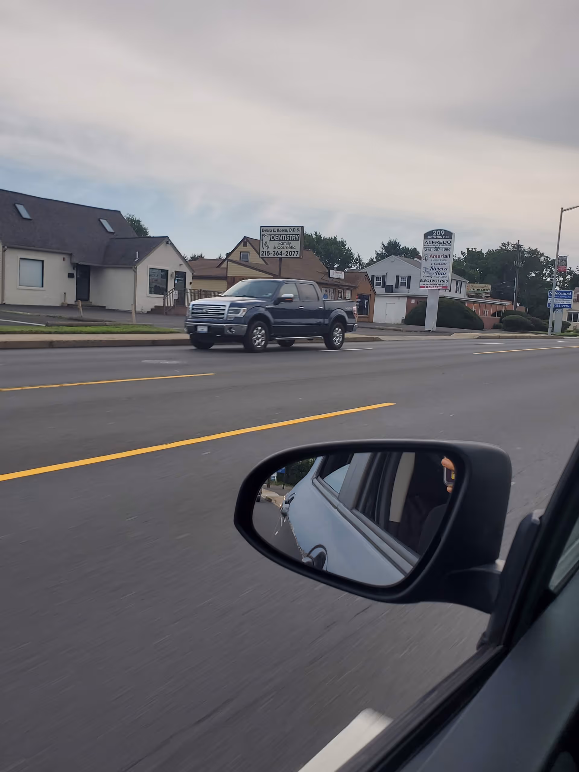 View from a moving car of storefronts and signs along a suburban street.