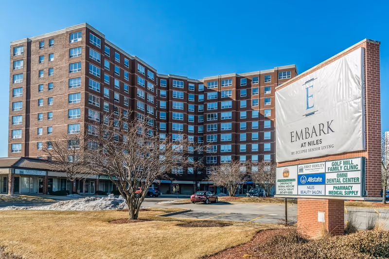 Multi-story brick senior living building front with a large 'Embark at Niles' sign and parking area under a clear blue sky.