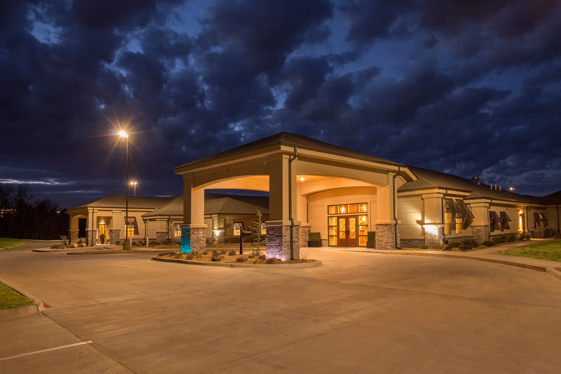A warmly lit senior living facility exterior at night with a covered entrance and driveway.