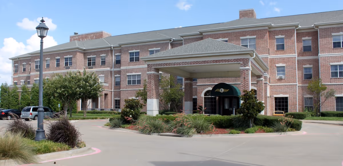 Exterior view of a three-story brick senior living facility building with a covered entrance, landscaped greenery, and a parking area with cars. The sky is partly cloudy.