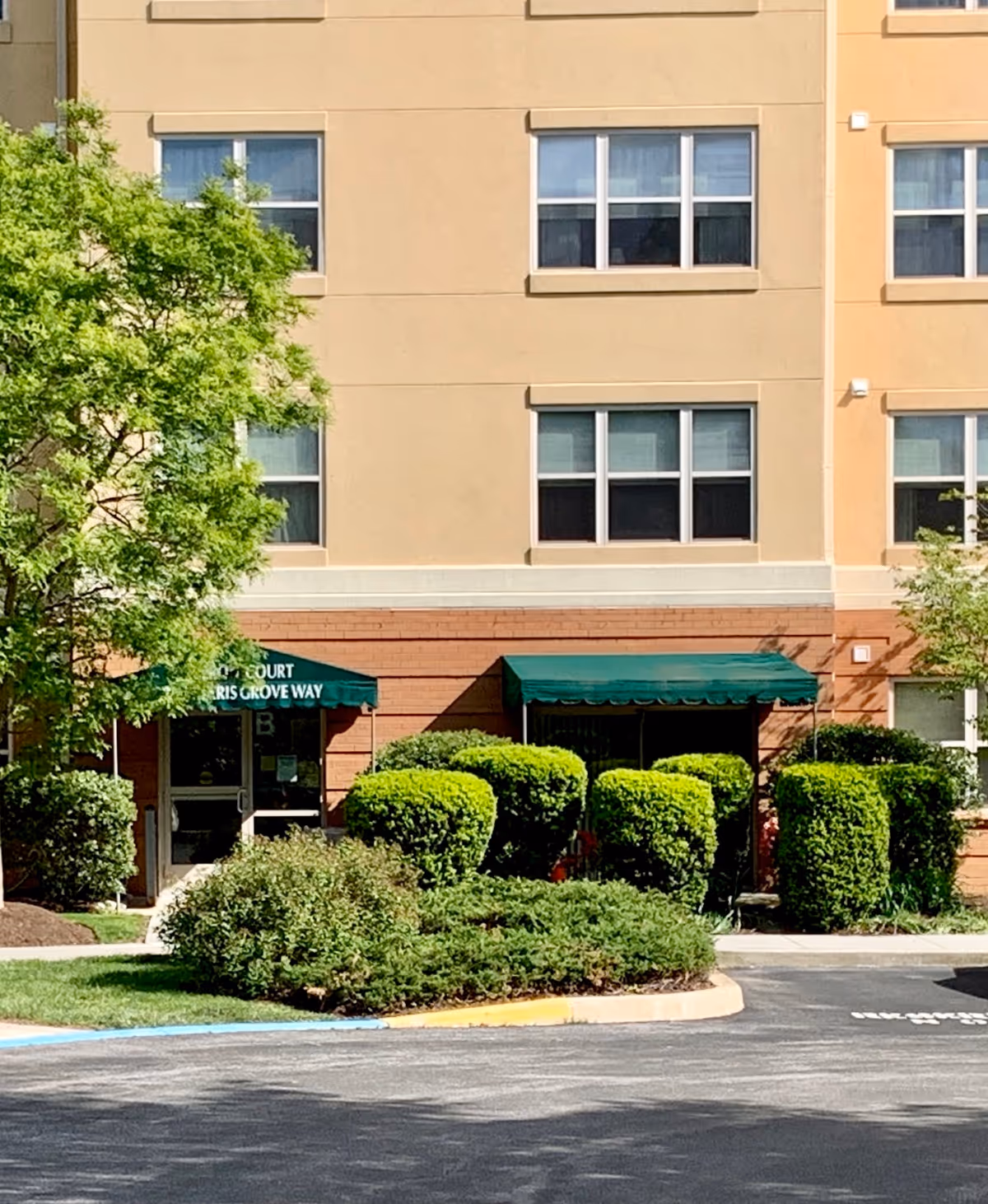 Beige multi-story senior living building with green awnings and trimmed shrubs at entrance.