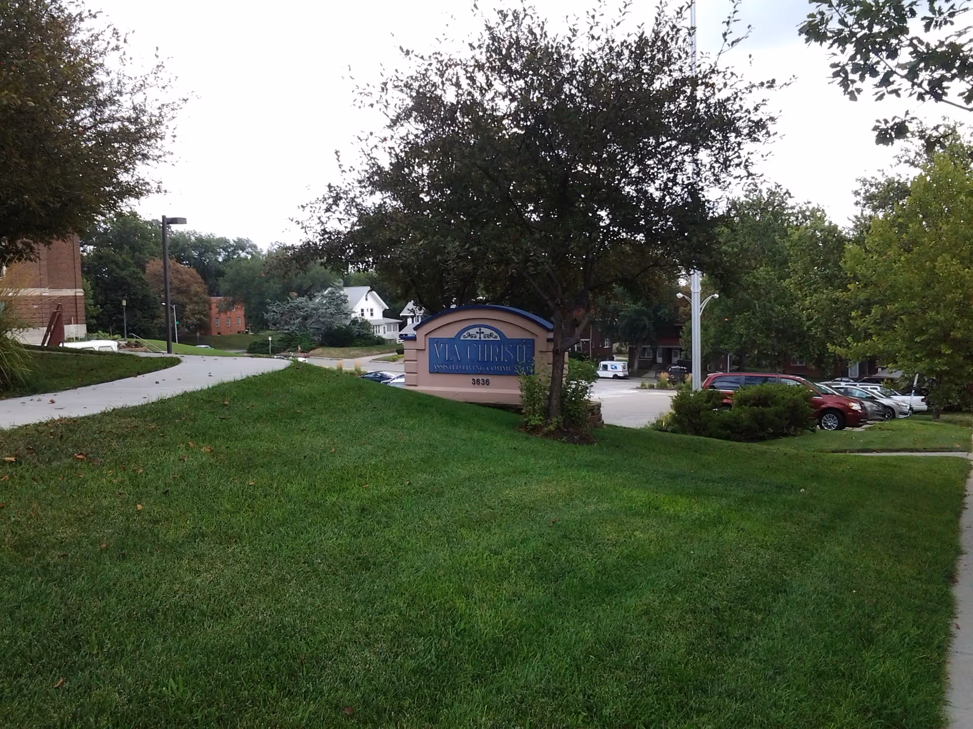 Exterior view of Via Christe Assisted Living sign, parking lot, and surrounding trees.