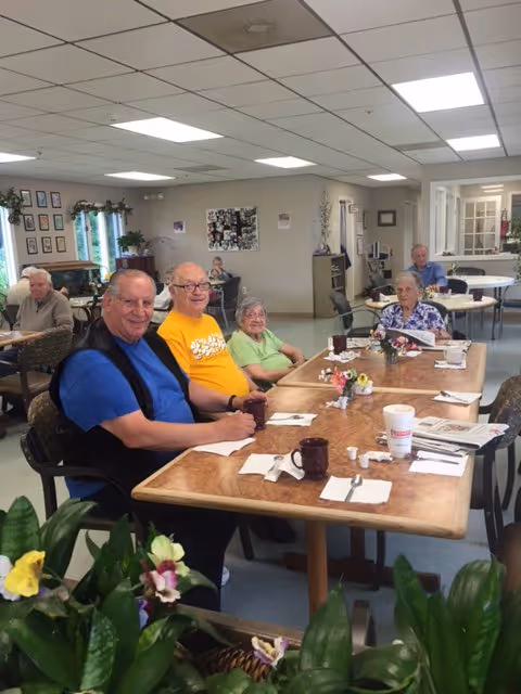 A group of elderly people seated at tables in a communal dining/activity room with plants in the foreground.