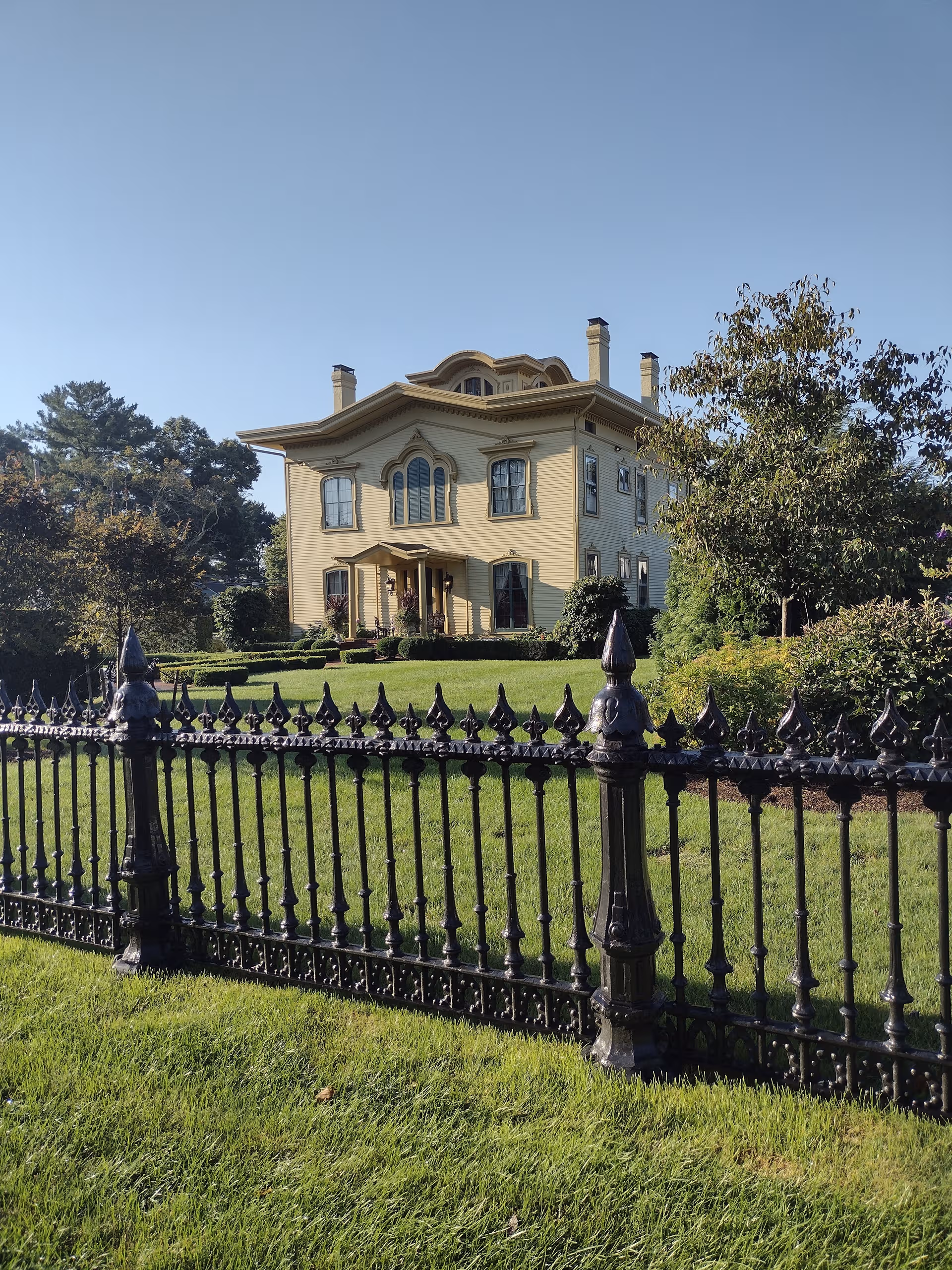 A large pale-yellow Victorian-style house with a black wrought-iron fence and manicured lawn.