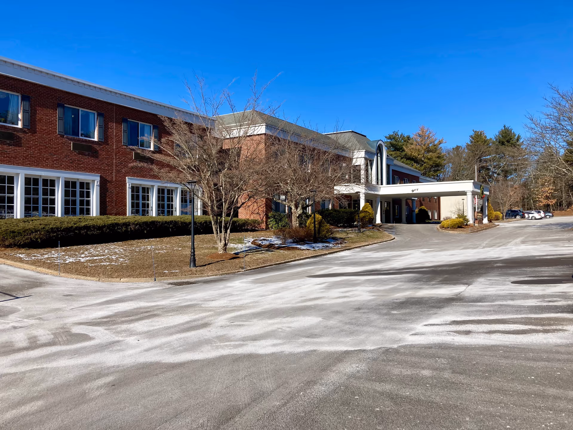Two-story red brick senior living building with a covered entrance/porte-cochere, driveway and sparse winter landscaping under a clear blue sky.