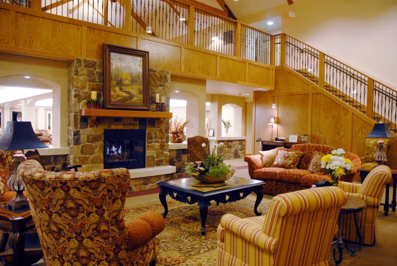 A cozy living room area in Pine Hills Retirement Community featuring a stone fireplace with a painting above it, surrounded by comfortable upholstered chairs and sofas in warm tones. A wooden coffee table with a plant centerpiece sits on a patterned rug. The room has wooden paneling and a staircase with wrought iron railings leading to an upper level.