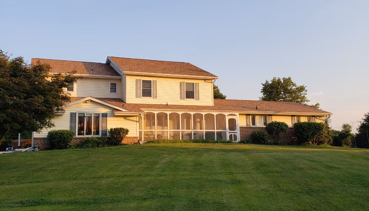 A two-story residential building with beige siding and a brown shingled roof, surrounded by green grass and bushes. The building features a screened-in porch and several windows, set against a clear sky during sunset.