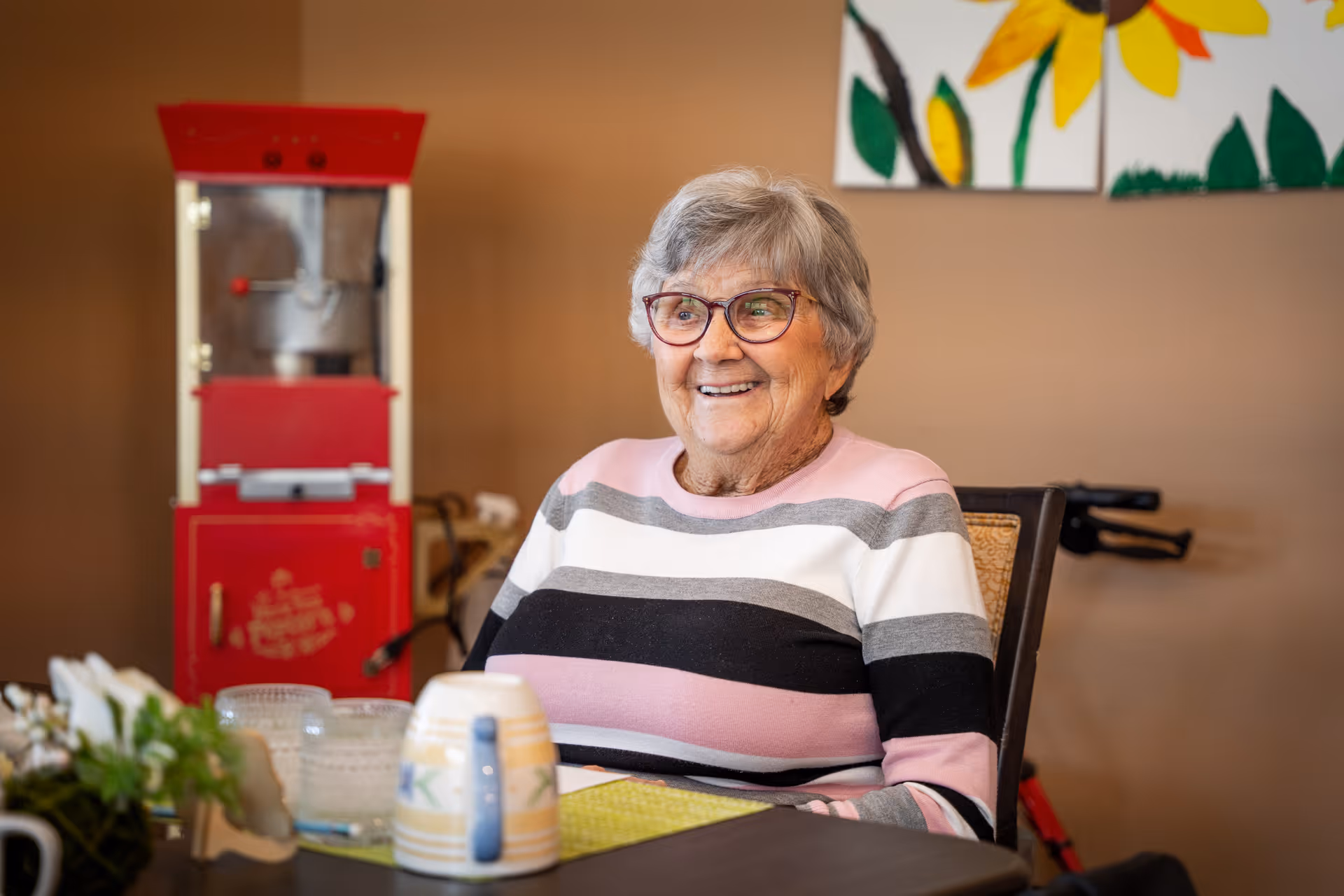 An elderly woman with gray hair and glasses wearing a striped sweater is sitting at a table in a room. On the table are a mug, some glassware, and a small plant. Behind her is a red popcorn machine and a wall with colorful artwork featuring flowers.