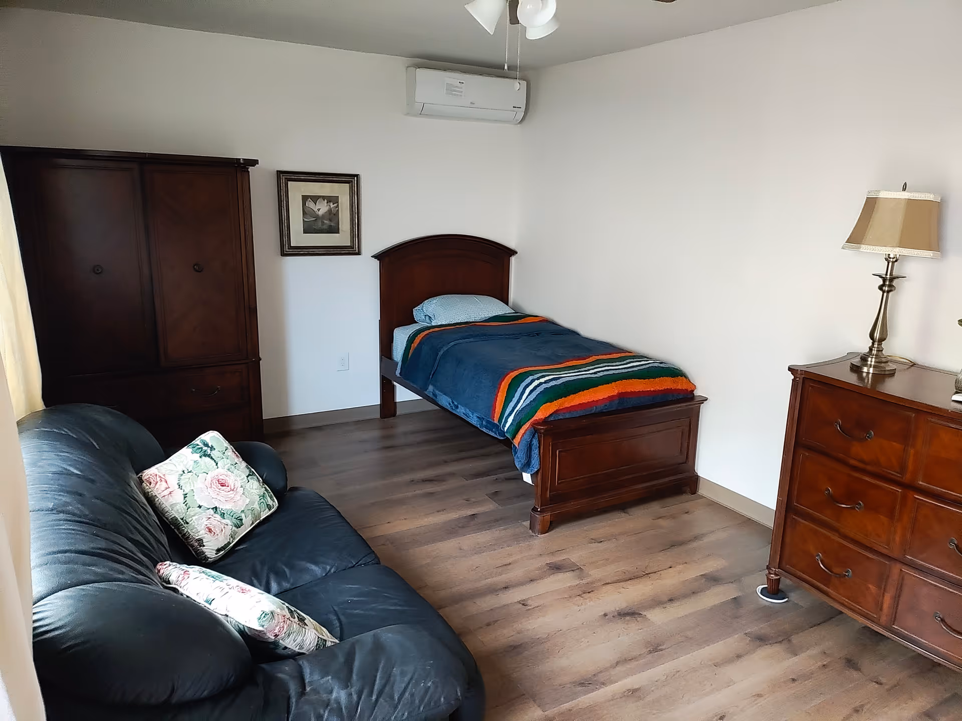 A furnished bedroom in a senior living facility with bed, sofa, dresser, and wardrobe.