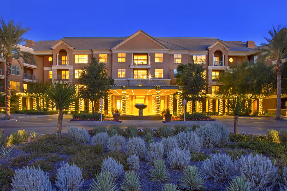 A warmly lit senior living facility entrance with stone facade and landscaped front garden at dusk.