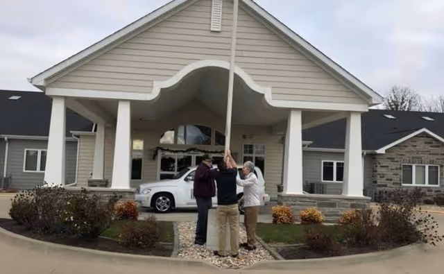 Three people stand under a covered entrance in front of a senior living facility.
