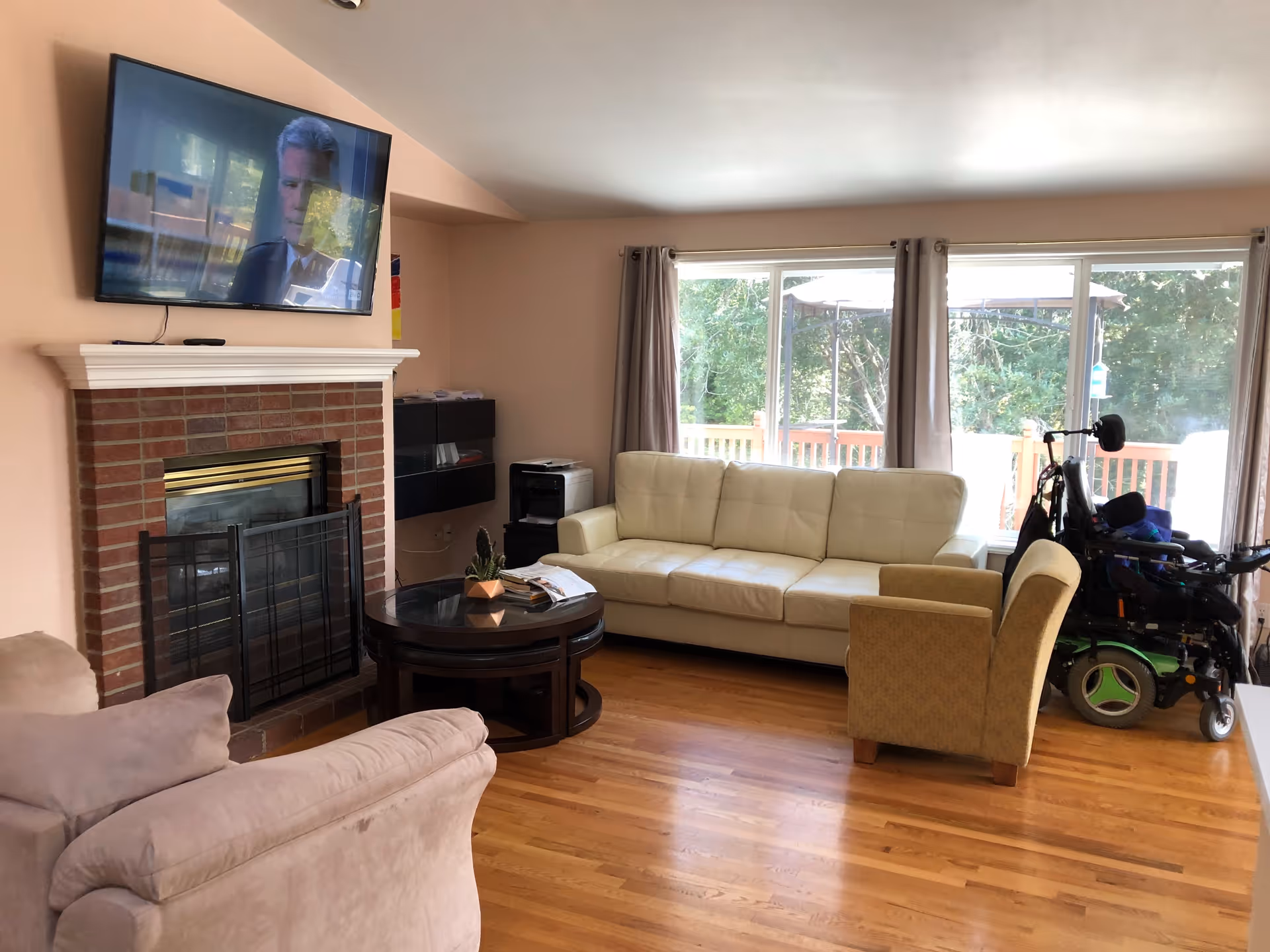Cozy living room with brick fireplace, white sofa, armchairs, coffee table, wheelchair, and mounted TV.