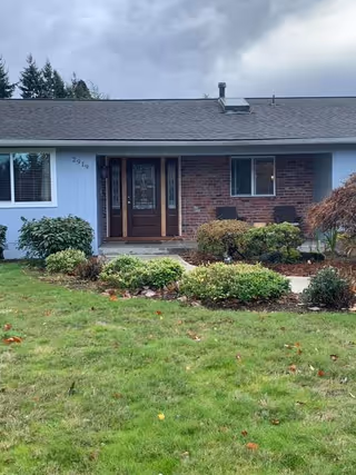 A single-story home with blue siding, a brick entry, and a landscaped front yard with shrubs and two chairs near the entrance.