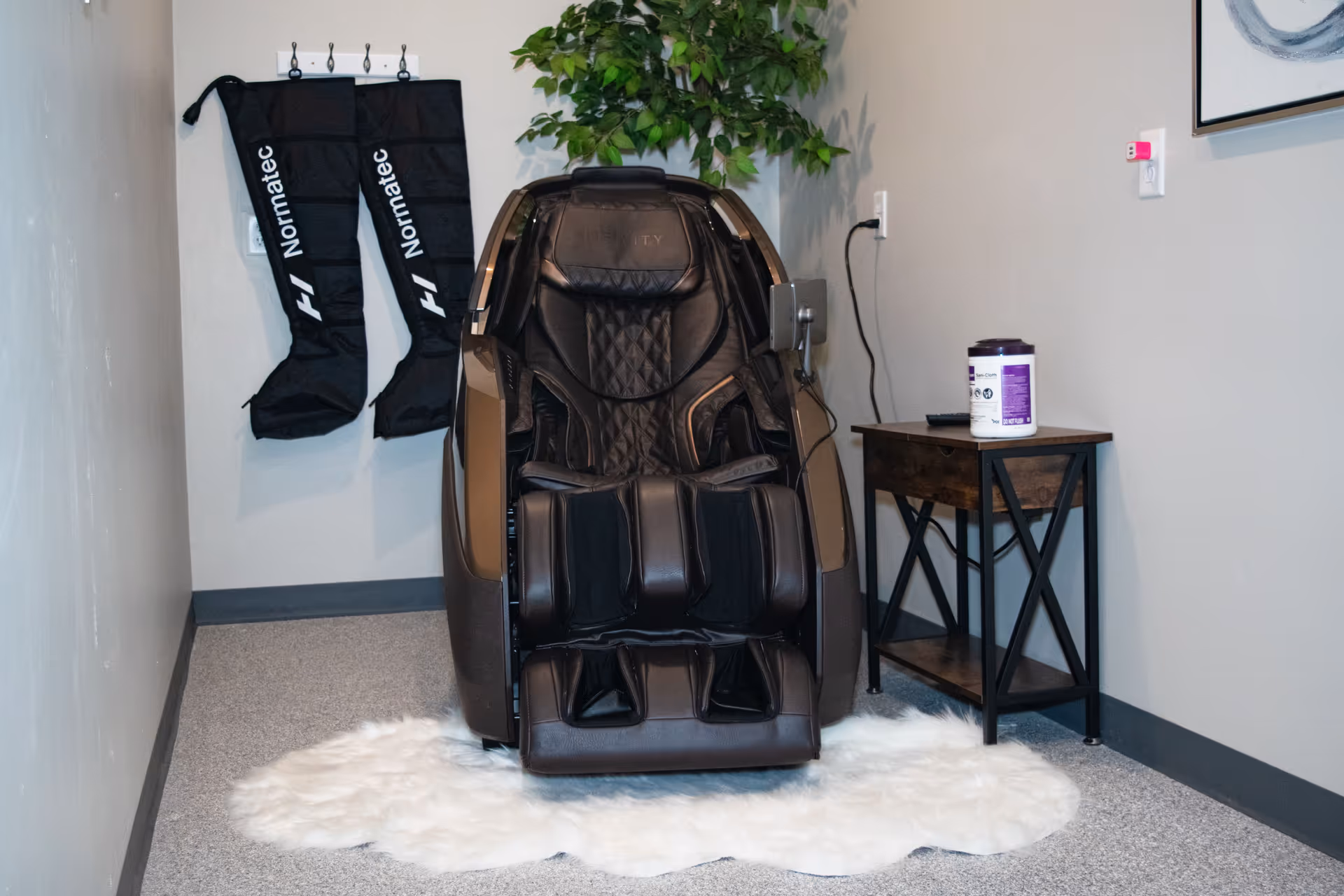 A black massage chair sits in a corner with a table, leg sleeves, and a rug.