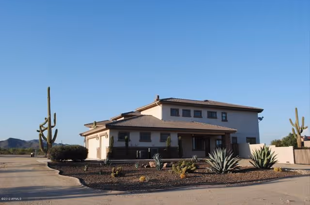 Exterior view of a two-story residential building with a tiled roof, surrounded by desert landscaping including cacti and shrubs, under a clear blue sky.