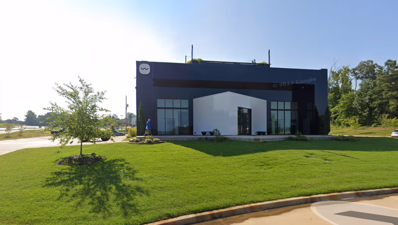 Modern dark building with white entry canopy, surrounding green lawn and a person nearby.